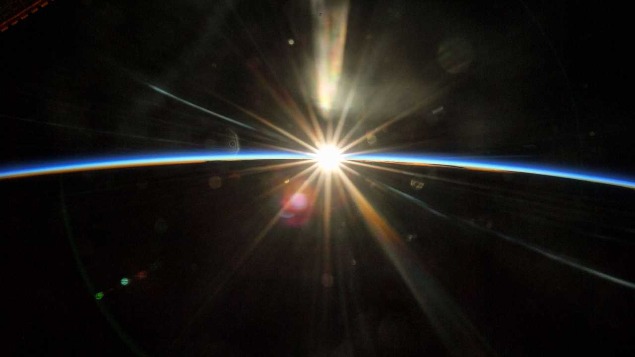 sun rising pierces blackness of space against the dark curvature of the earth with thin blue line of daytime atmosphere on the horizon