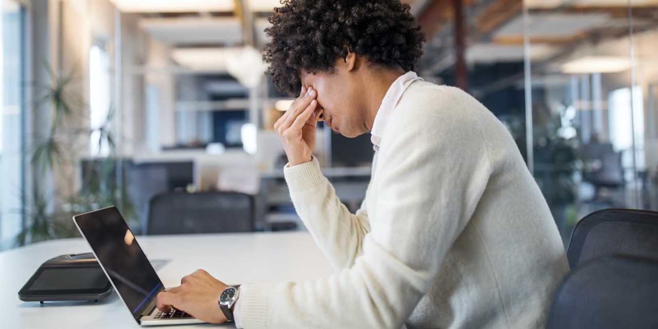 A person working in an office and looking stressed. He is working at a table with his laptop.