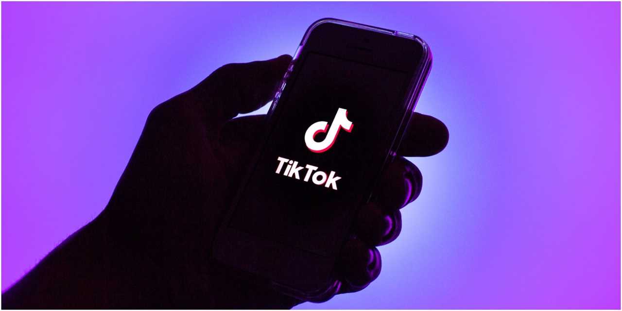 Hand holding a phone with a TikTok logo on the screen. Background is purple.