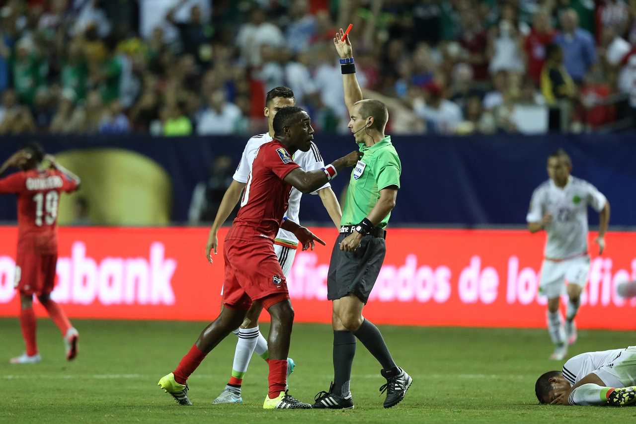 A soccer referees holds a red card up while a player jabs him in the chest, with another player on the ground to the right