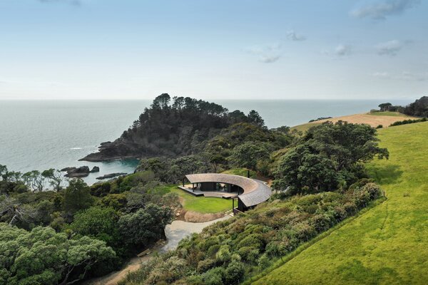 "Curled into a natural amphitheatre, this gentle timber house turns to face the sun rather than the sea. Responding to the landform, its simple semicircle creates north-facing spaces sheltered from onshore winds. One corner braves the sea cliff, facing the salt and spray, but most of the house is surrounded by bush and orients towards a small valley—the ocean all the more present for being ignored formally."