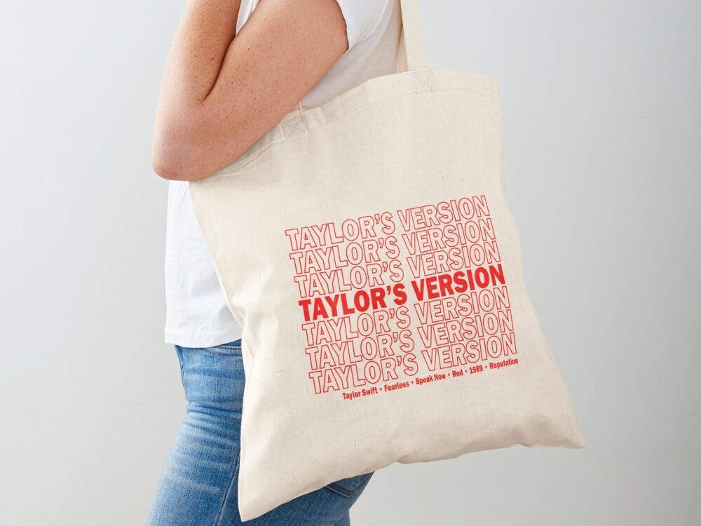 Person holding a canvas tote bag from Redbubble that says "Taylor's Version" in repeated red font, one of the best gifts for a Taylor Swift fan