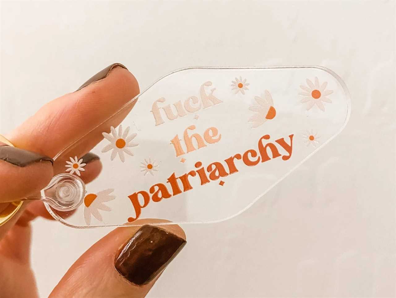 a hand holding a clear keychain from Etsy with "fuck the patriarchy" and flowers printed on it, one of the best gifts for a Taylor Swift fan