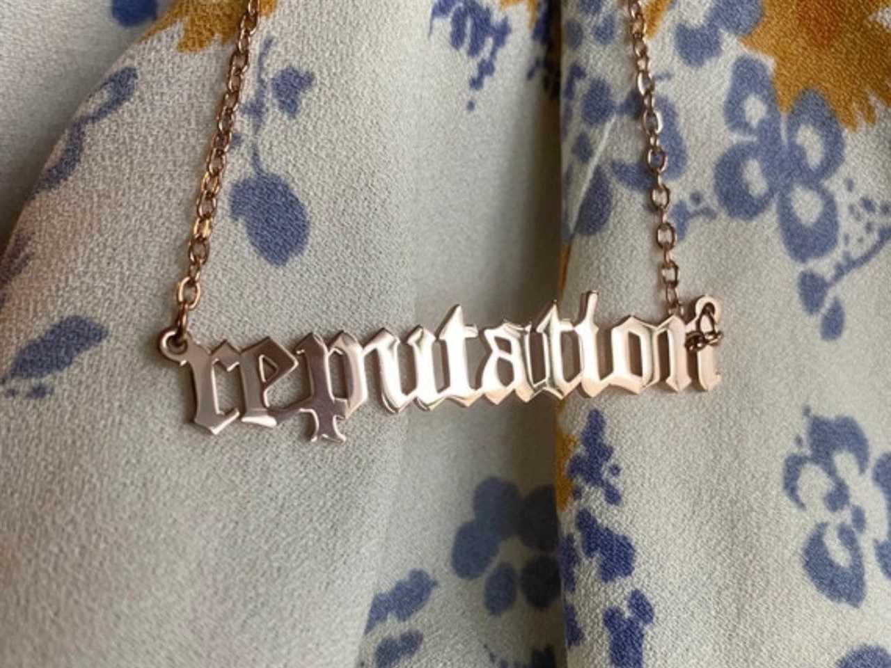 A gold pendant necklace of Taylor Swift's "Reputation" album title from Etsy placed on a floral blanket, one of the best gifts for a Taylor Swift fan