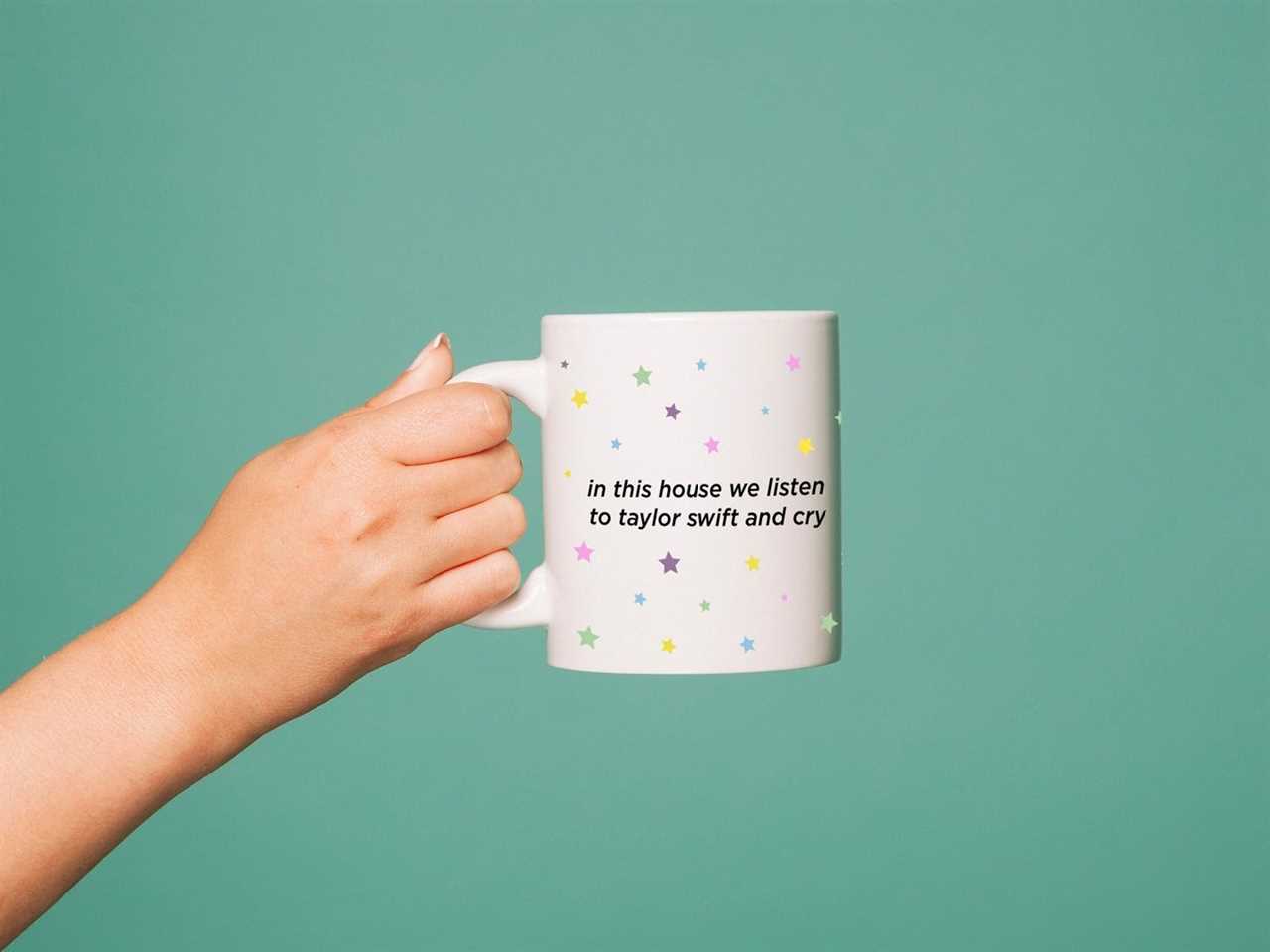 Person's hand holding a white mug printed with various colors of stars and text that says "in this house we listen to Taylor Swift and cry" from Etsy, one of the best gifts for a Taylor Swift fan