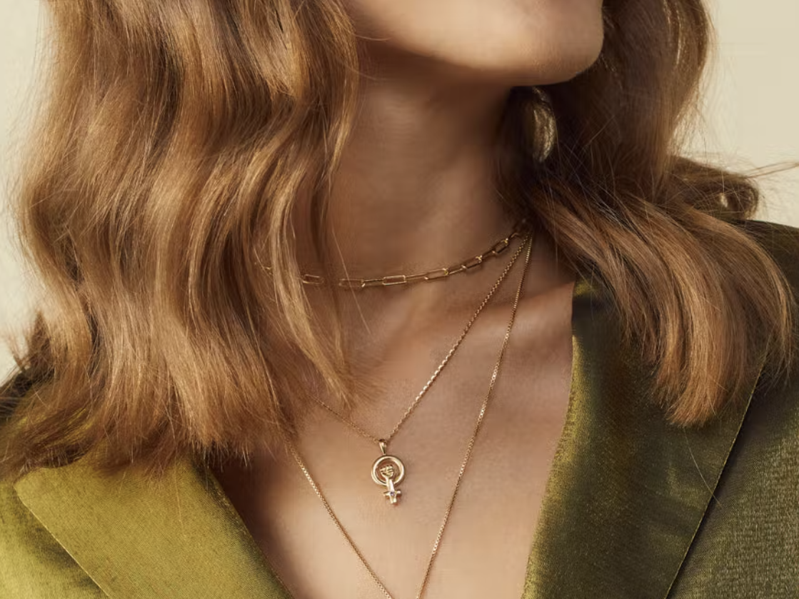 a person wearing a gold Awe Inspired "woman power" necklace with a pendant in the shape of a female sex symbol with a raised fist in the center, one of the best gifts for a Taylor Swift fan