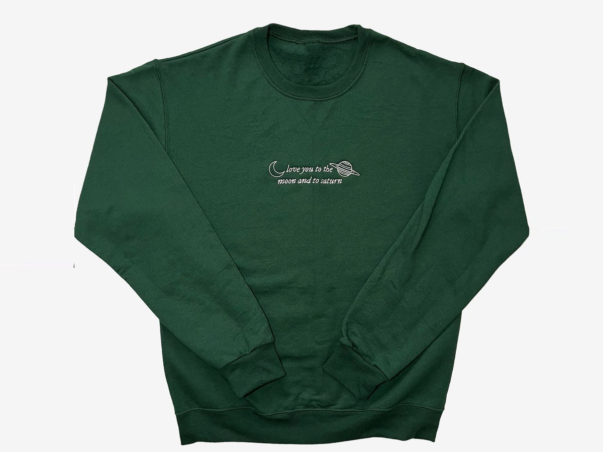 A forest green sweatshirt from Etsy that is embroidered with text that says "love you to the moon and to Saturn," lyrics from Taylor Swift's song "Seven", one of the best gifts for a Taylor Swift fan