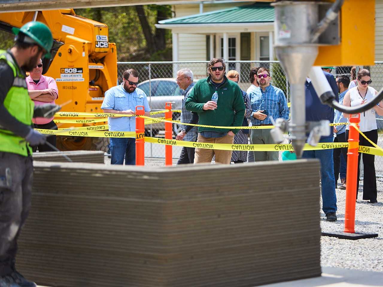 A 3D printer printing concrete onto a wall. There's groups of people next to the printer looking on.