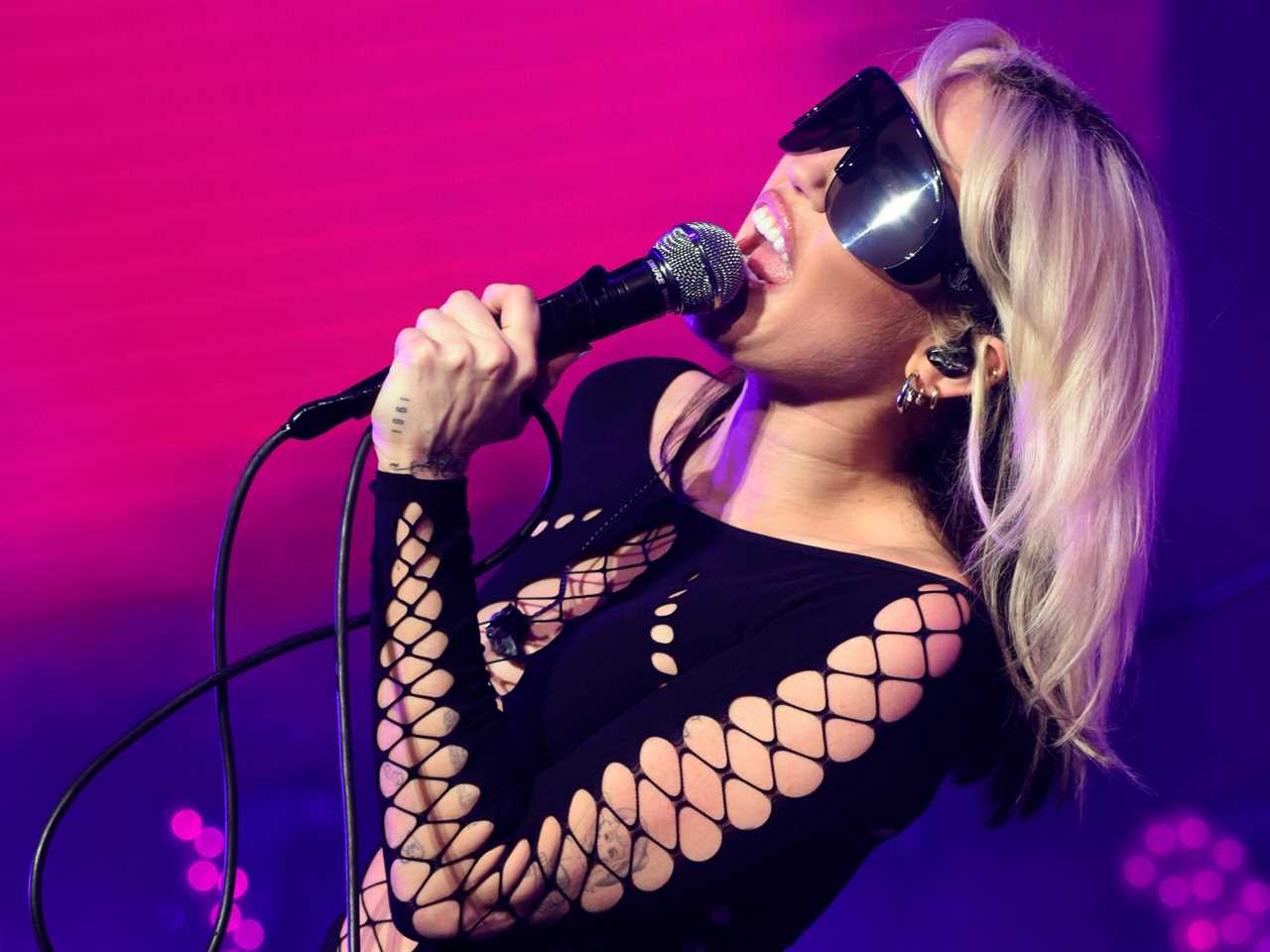 Miley Cyrus sings into a microphone with sunglasses on in front of a pink and purple backdrop.