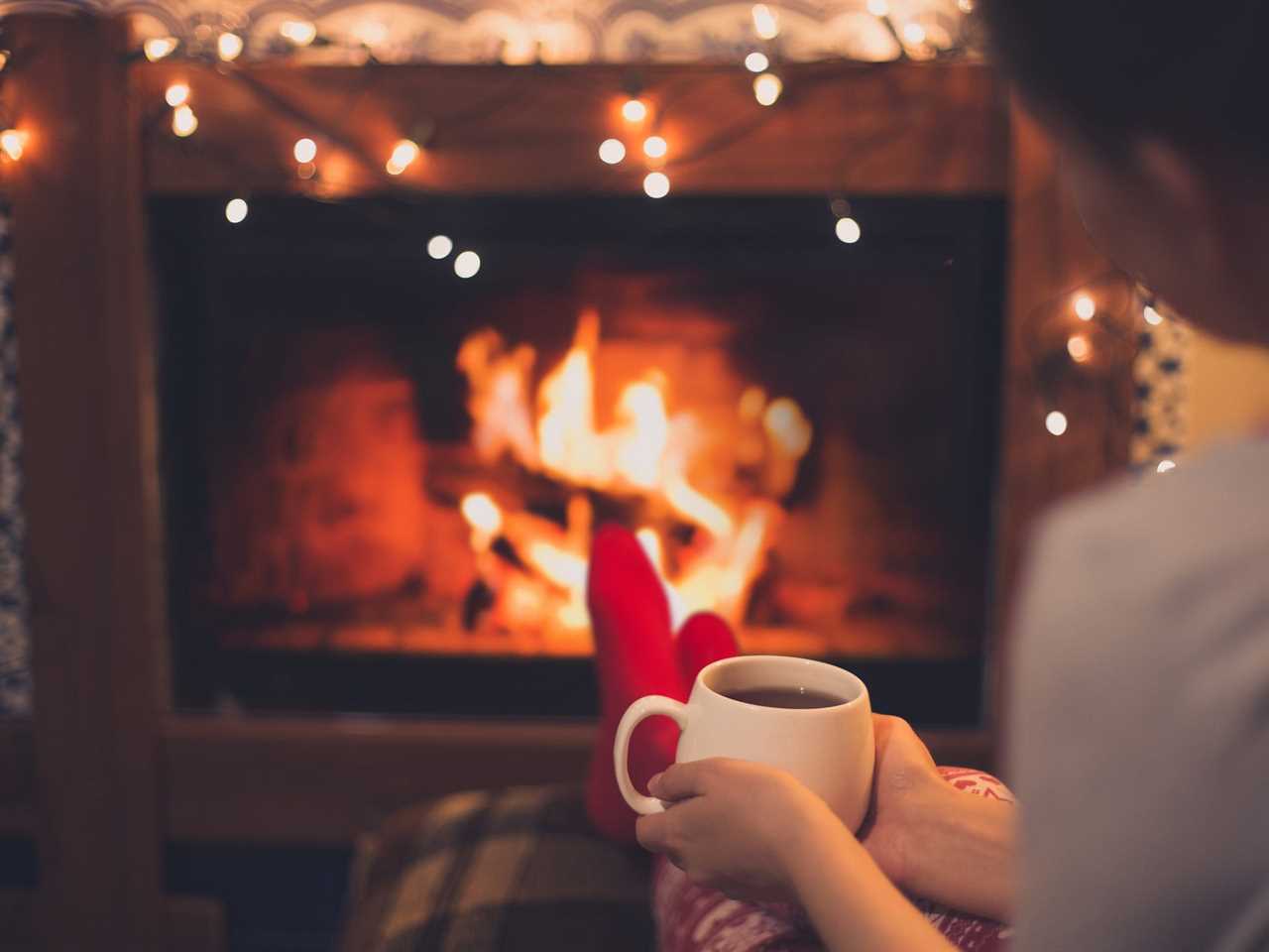 Close up cup of hot tea in woman's hands sitting near fireplace with festive Christmas lights in cozy room.