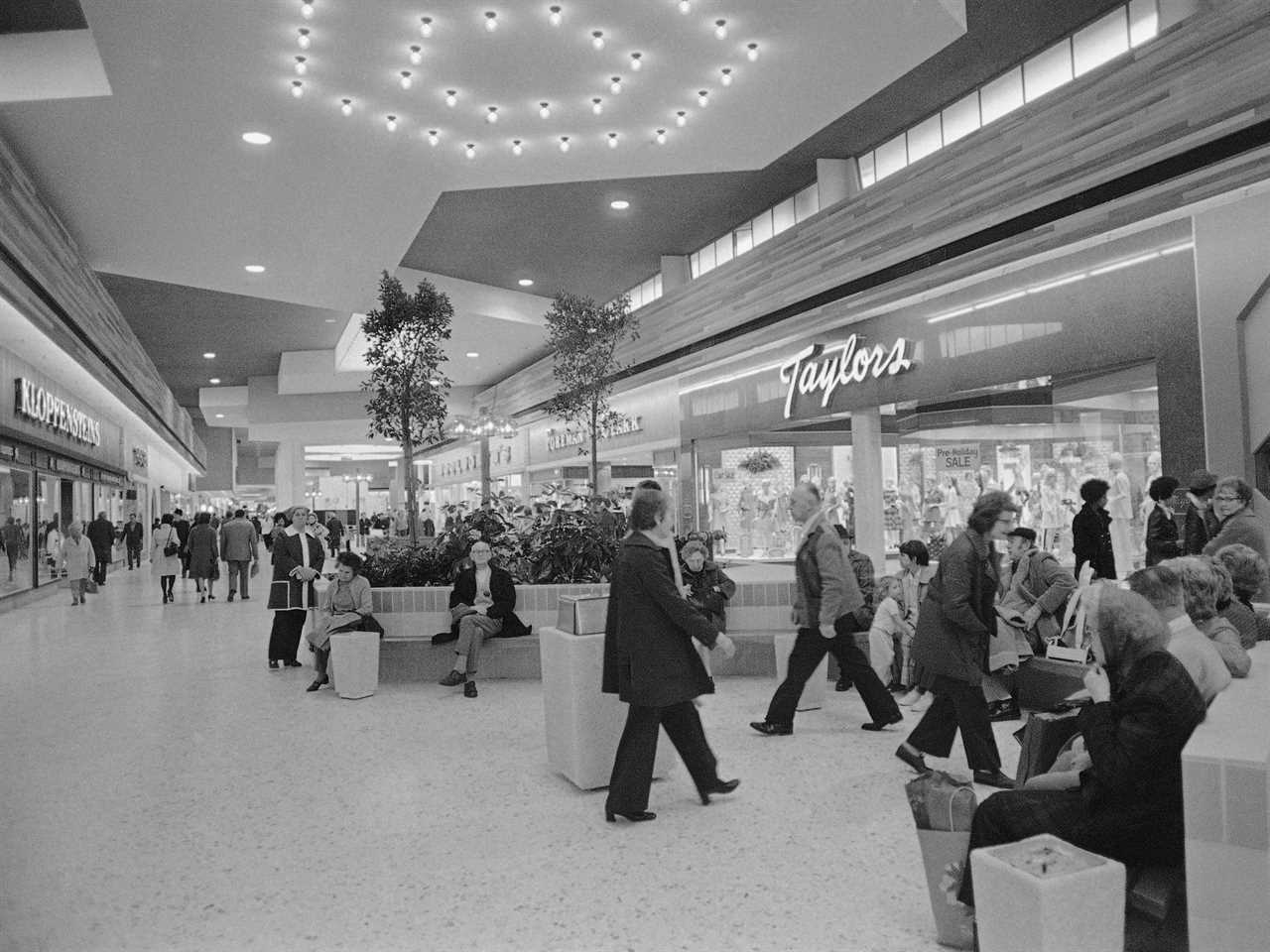 Interior shot of a shopping mall in 1974 with shoppers walking around and sitting on benches