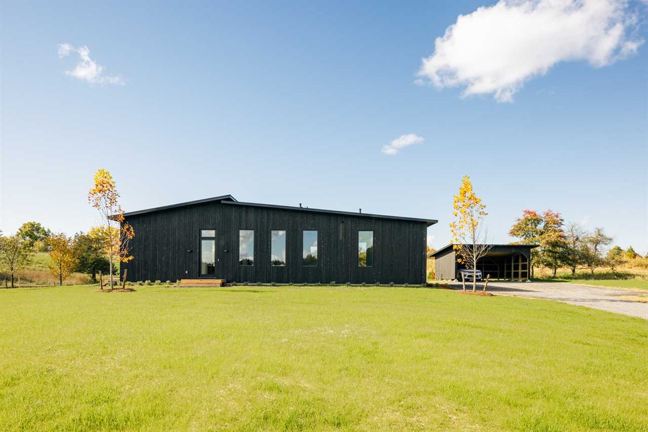 The black-stained cedar facade pops against the home's sprawling lawn and surrounding countryside. A detached carport sporting a similar style offers additional storage.