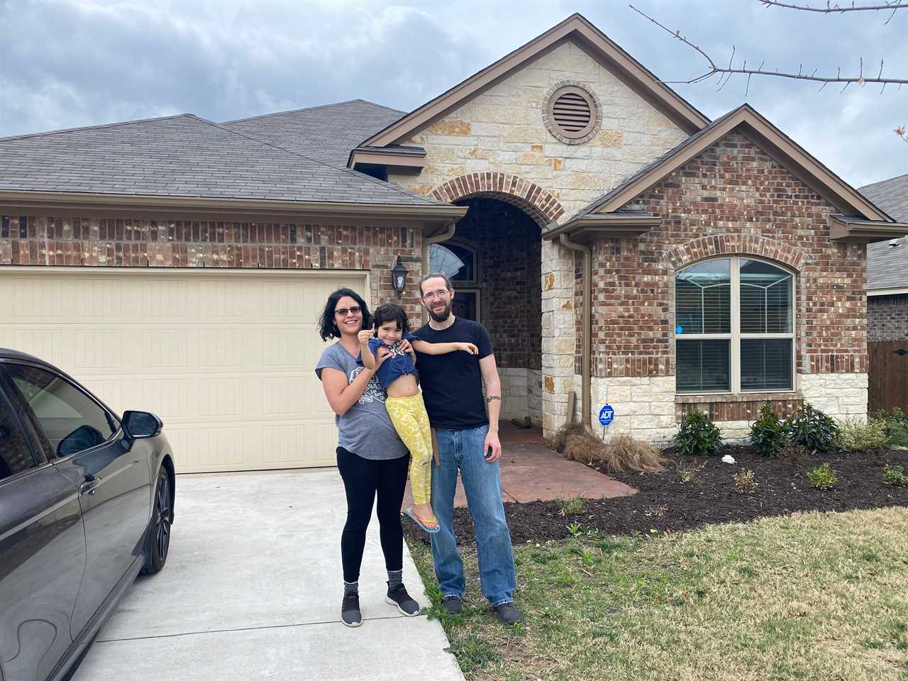 Mark Sorvillo, in a black tee shirt and jeans stands beside his wife who lifts up their daughter. All are smiling in front of their new house, with a brown brick facade and a garage in New Braunsfel, Texas