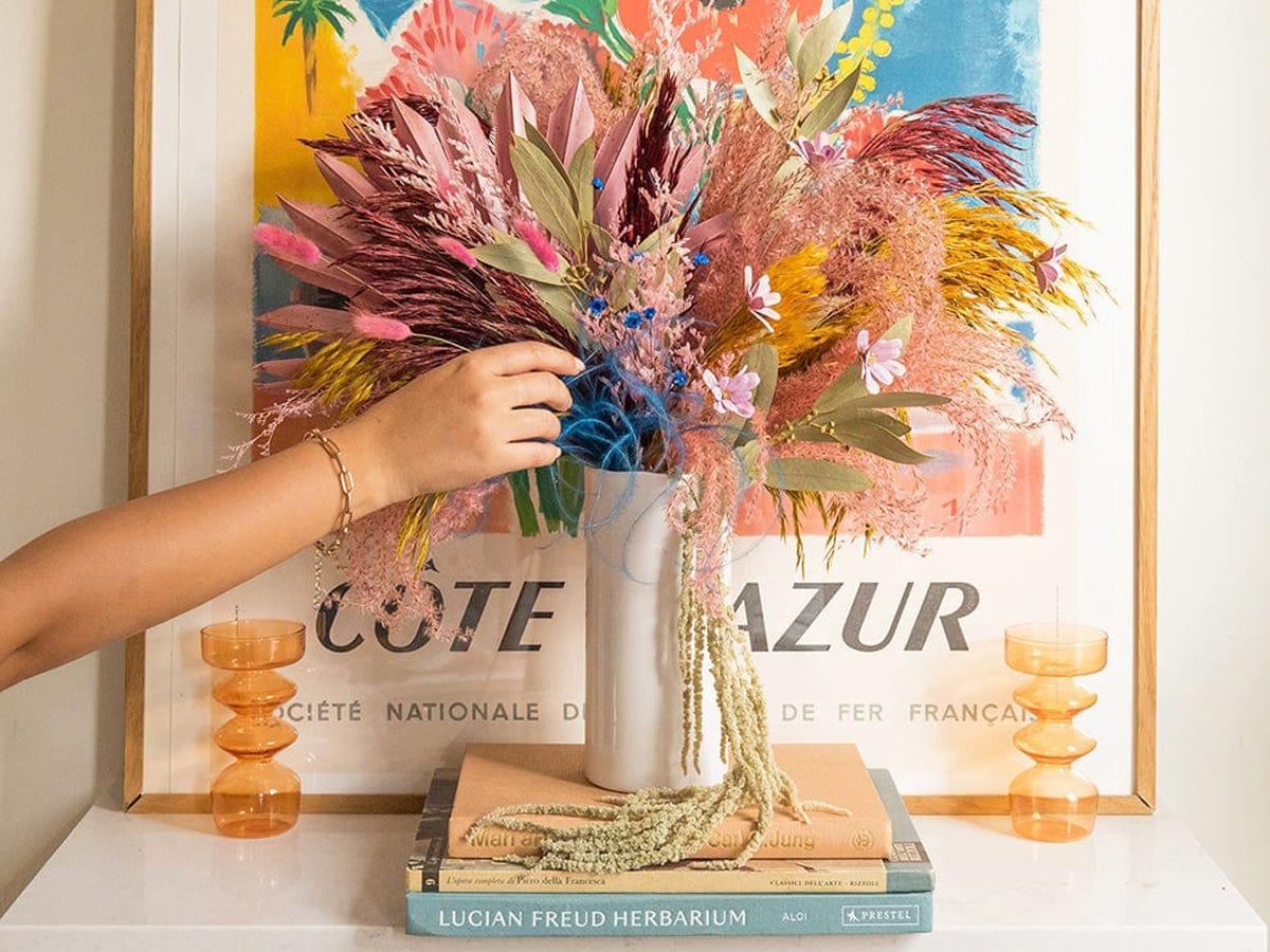 Hand reaching towards a colorful bouquet of dried flowers set on top of a stack of books in front of framed artwork.