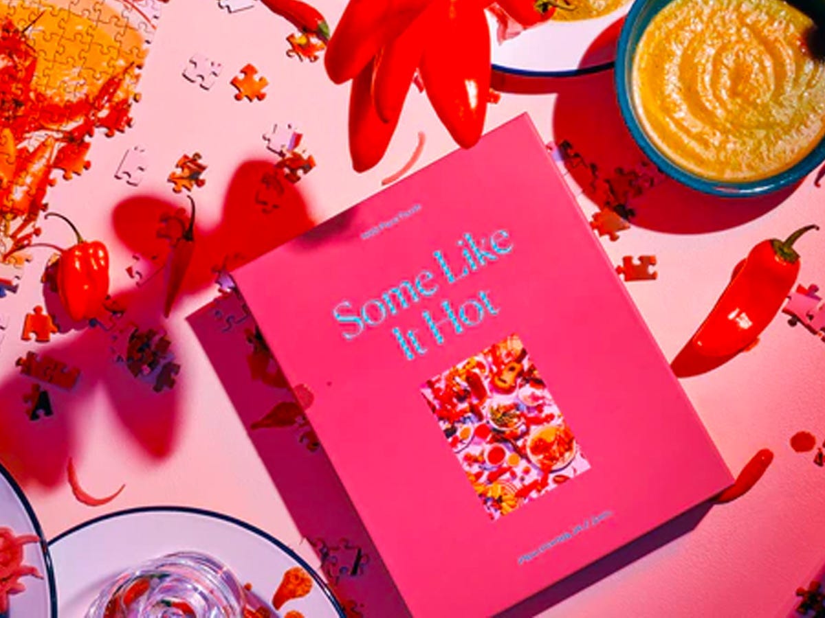 Pink box containing the Some Like It Hot puzzle on a table surrounded by puzzle pieces, hot peppers, and a bowl of salsa.