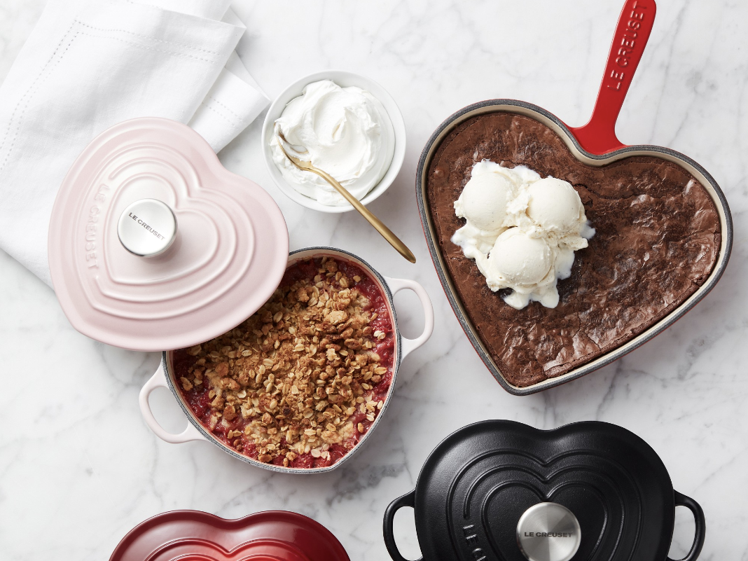 A few heart-shaped Le Creuset cast iron pans in pink, red, and black set out on a marble countertop. The red skillet holds a brownie with ice cream scooped on top, the pink Dutch oven has the lid removed to show a fruit cobbler inside.