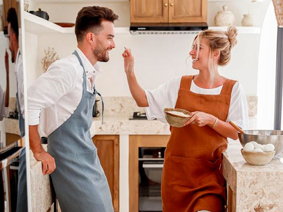 A couple leaning against counters in a kitchen both wearing different colors of the Magic Linen Pinafore Cross-Back Linen Apron