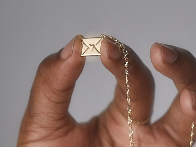 A hand holding up a gold catbird necklace featuring the smallest love letter charm.