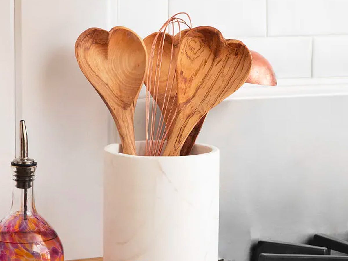 Three wooden heart-shaped serving spoons, a whisk, and a ladle in a utensil holder set on a kitchen counter next to a stove and a bottle of cooking oil.