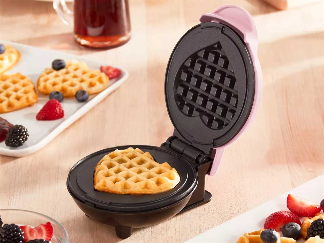 Dash mini waffle maker open to show a heart-shaped waffle inside, set on a table next to a plate of waffles and fruit.