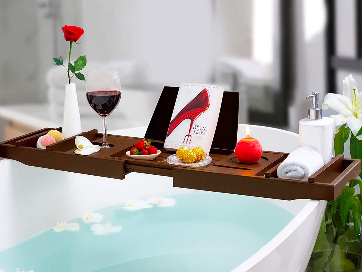 Royal Craft Wood Luxury Bathtub Caddy Tray set across a filled bathtub with a glass of wine, flowers, candle, strawberries, chocolate, and a copy of the "Devil Wears Prada" book on top of it.