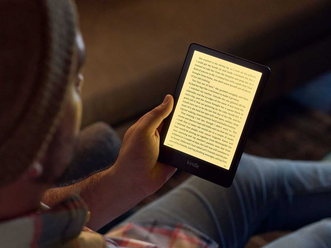 A person sitting on a couch while reading a book on an Amazon Kindle Paperwhite in low lighting.