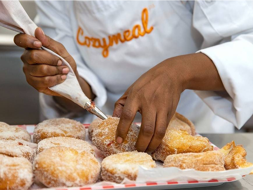 A person wearing a Cozymeal apron using a pipping bag to fill donuts with jelly.