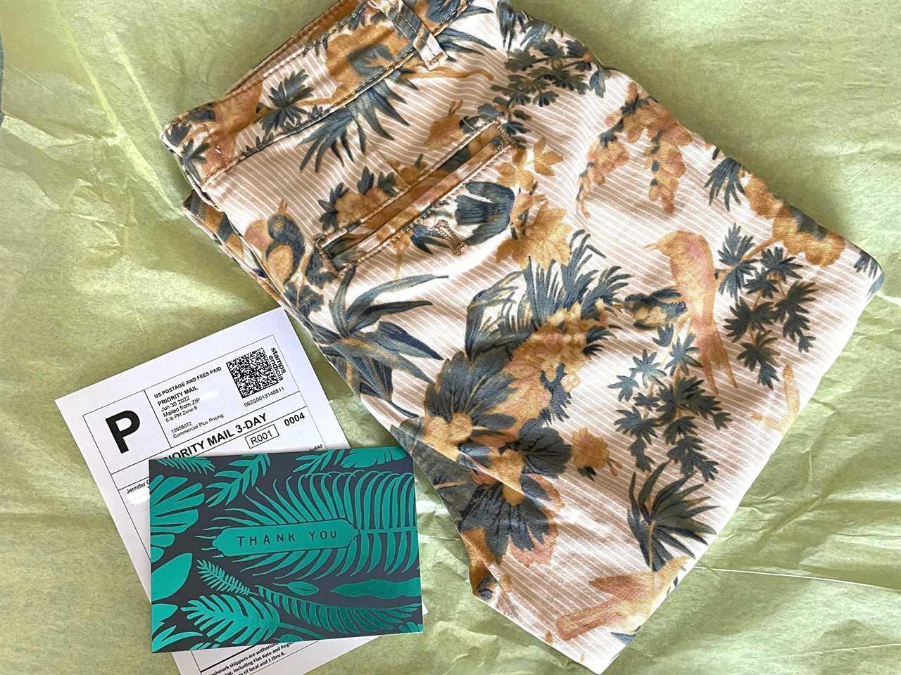 A pair of floral pants lays folded on top of yellow tissue paper next to a shipping label and thank you card.