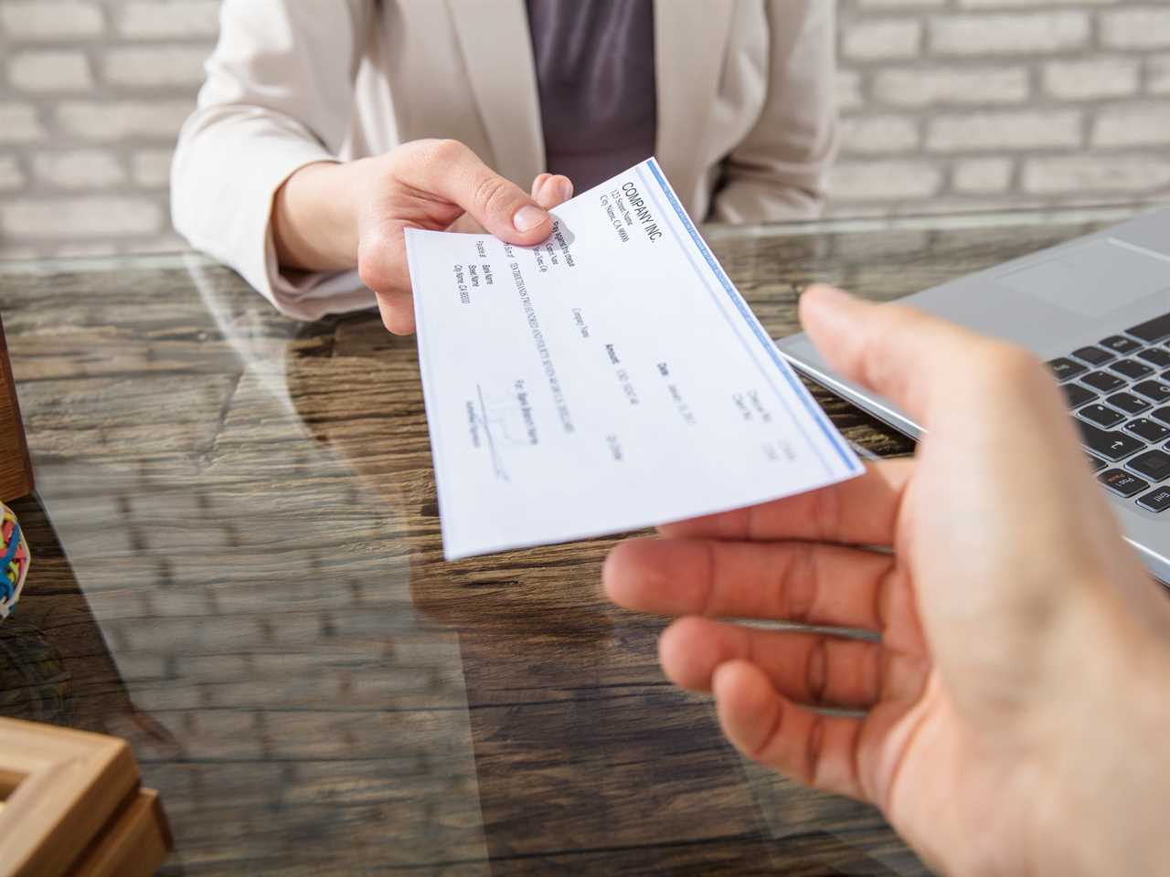 A person across a desk handing over a paycheck to a person with an outstretched hand.