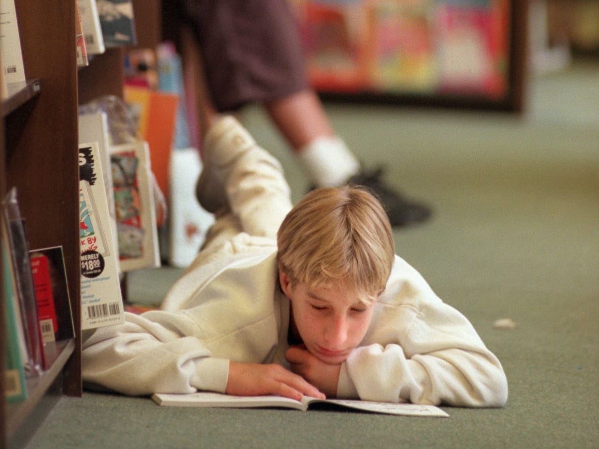 Teenage boy lays on stomach reading a book in front of book shelves at Barnes & Noble store