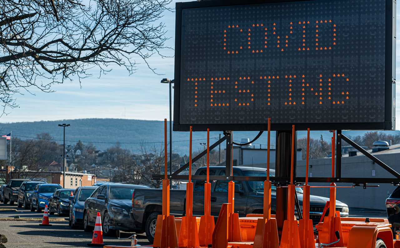 A line of cars near a light-up traffic warning-type sign reading "COVID testing," surrounded by orange construction cones