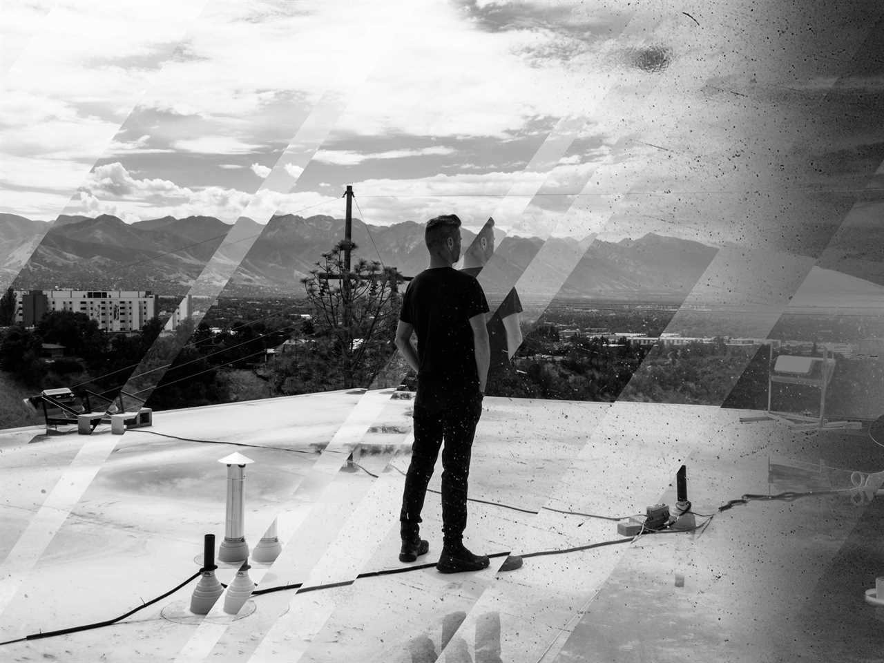 Black and white image of a man in a black T-shirt, standing on a roof and facing away from the camera toward a mountain vista.