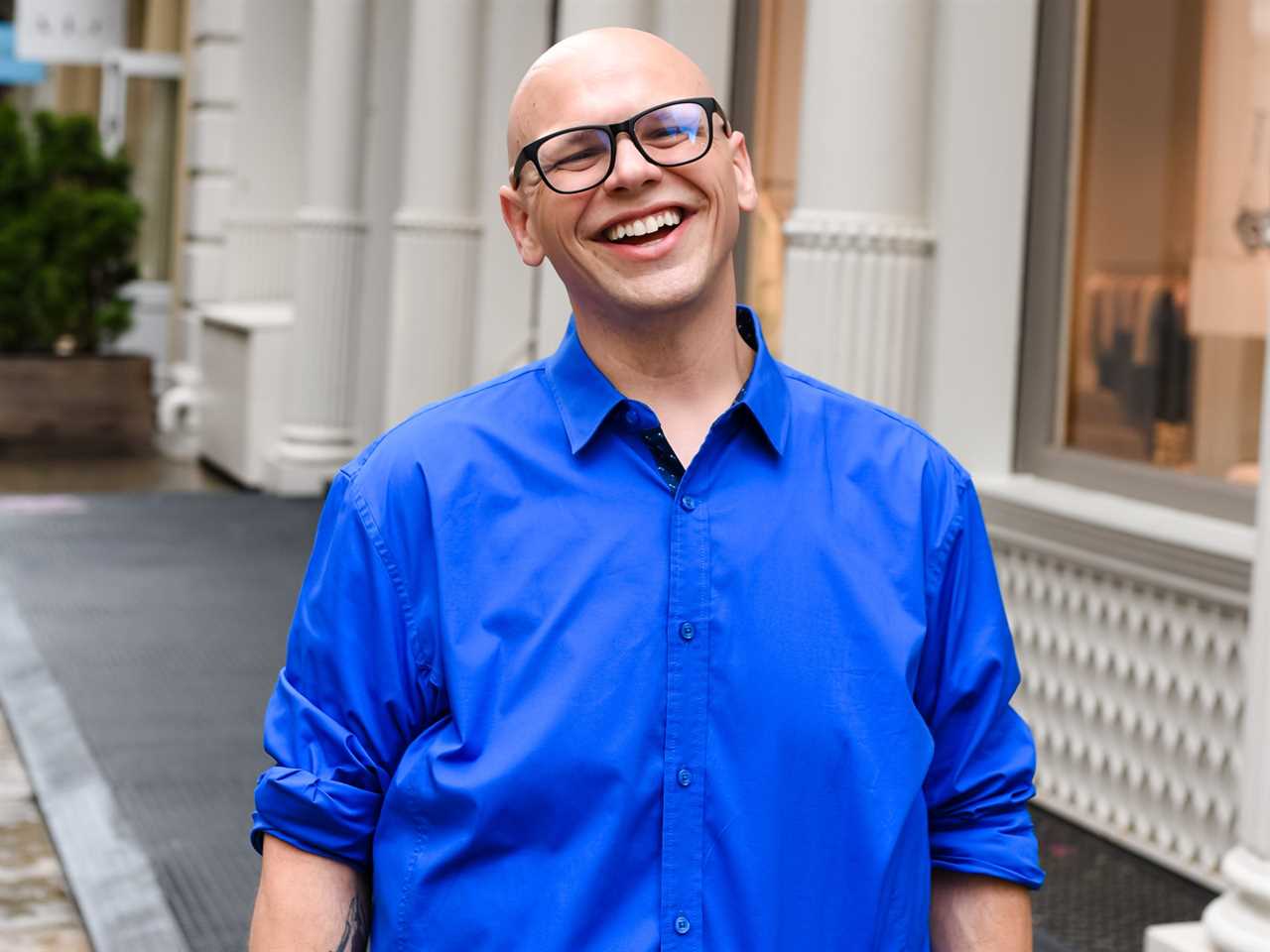 A photo of Kevin Dulaney smiling and wearing a blue button up, jeans, and a brown belt while walking on a sidewalk.