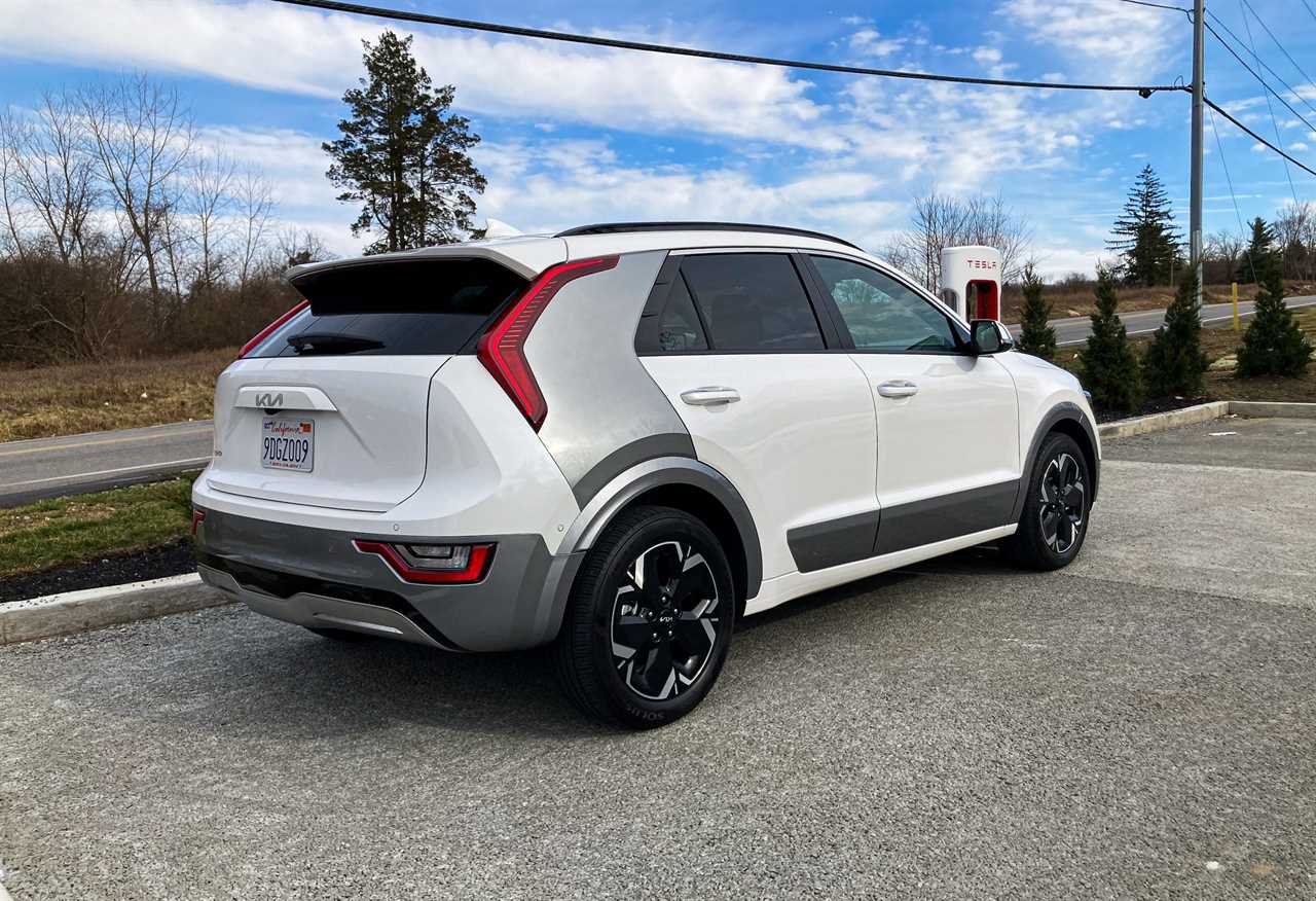 A white Kia Niro EV electric SUV seen from the rear in a parking lot against a blue sky.