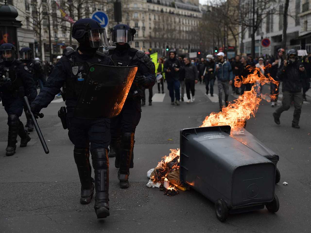 Riot police walk past a garbage can set on fire by demonstrators during a protest