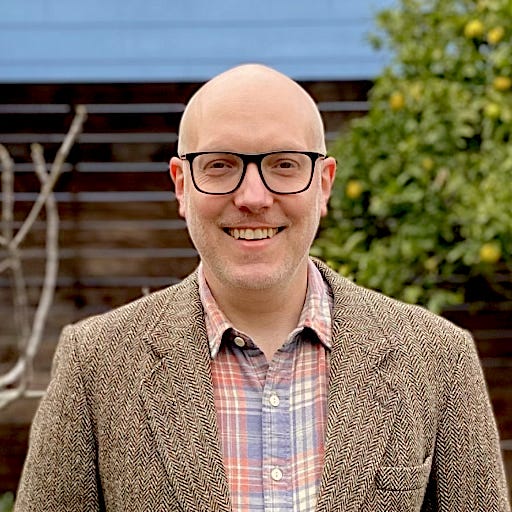 Headshot of Nick Reavill who's wearing a textured blazer over a plaid shirt.