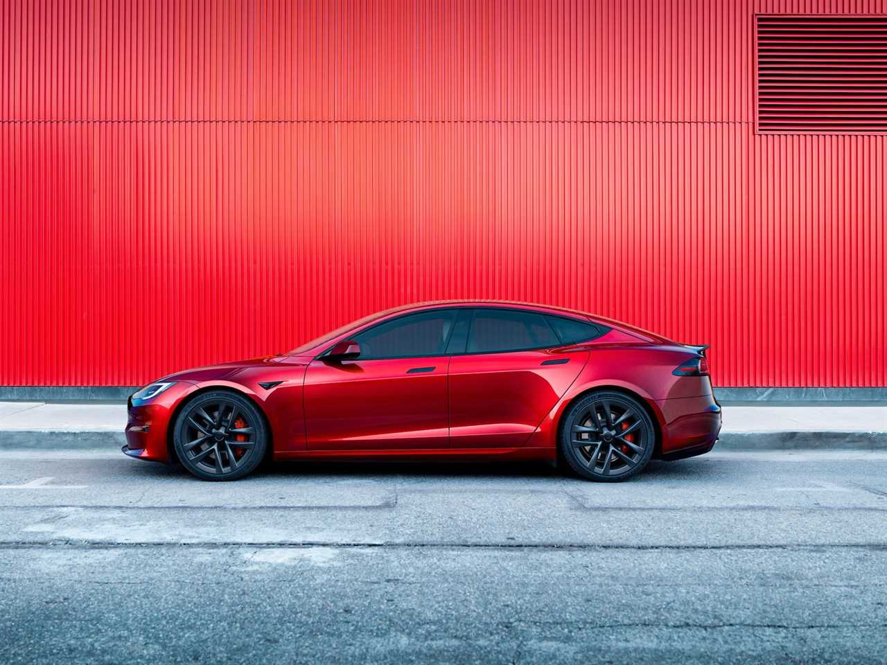 A red Tesla Model S. parked on a paved road against a red building.