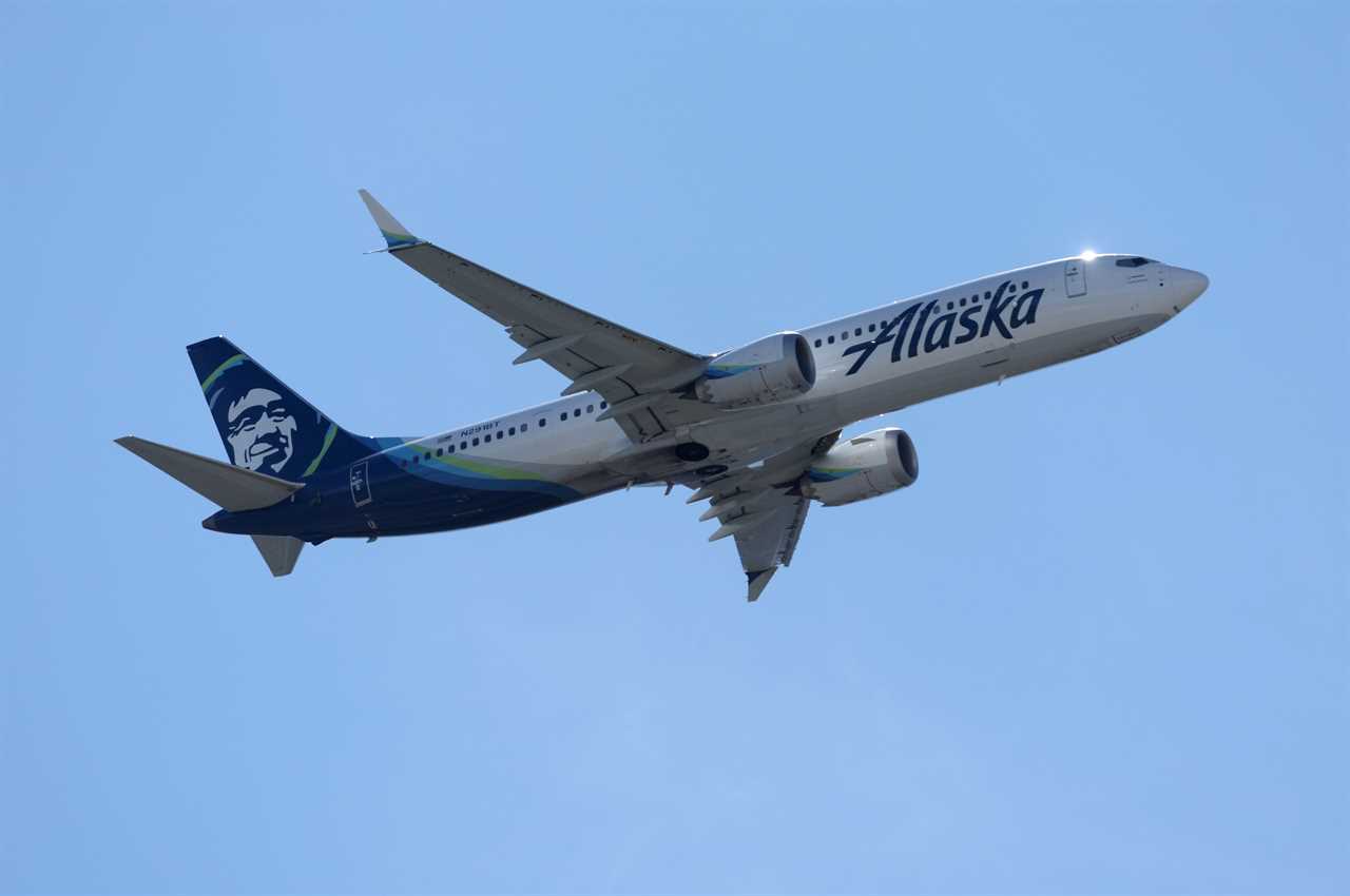 A white and blue Alaska Airlines plane in midair, set against a blue sky.