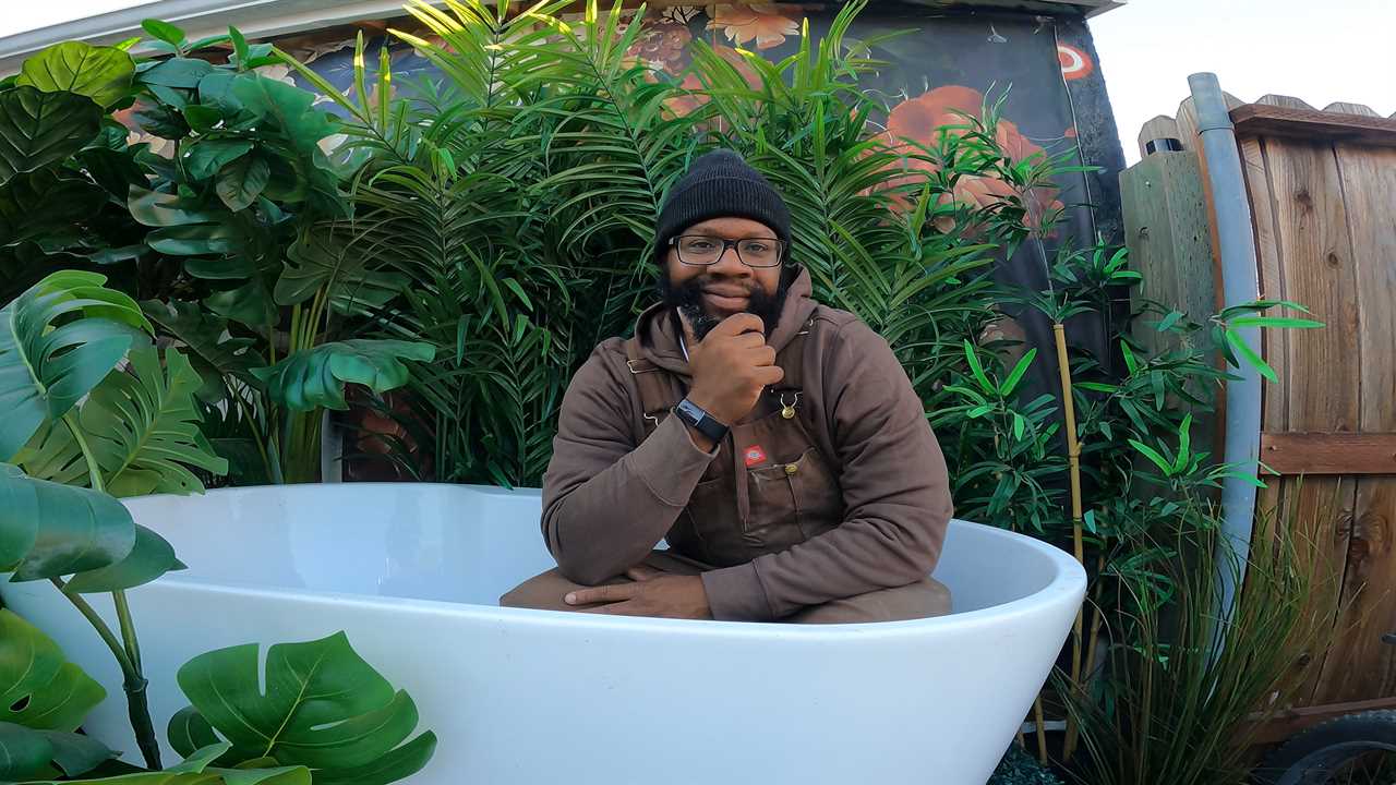 Ansel Troy posing for a picture in a bathtub surrounded by plants outside his tiny home.