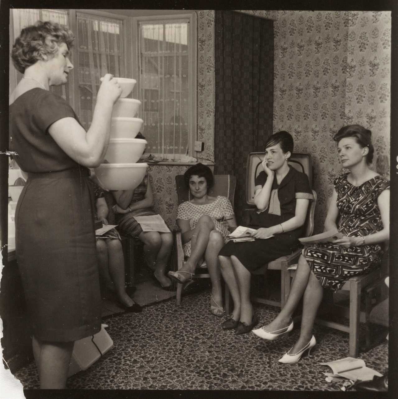 A woman holds five Tupperware bowls stacked on top of each other while she addresses other women in a home in the UK in 1963.