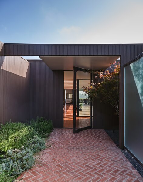 A wide brick patio leads to the main entrance, where a black-framed glass door opens to the sprawling, sunbathed interiors.