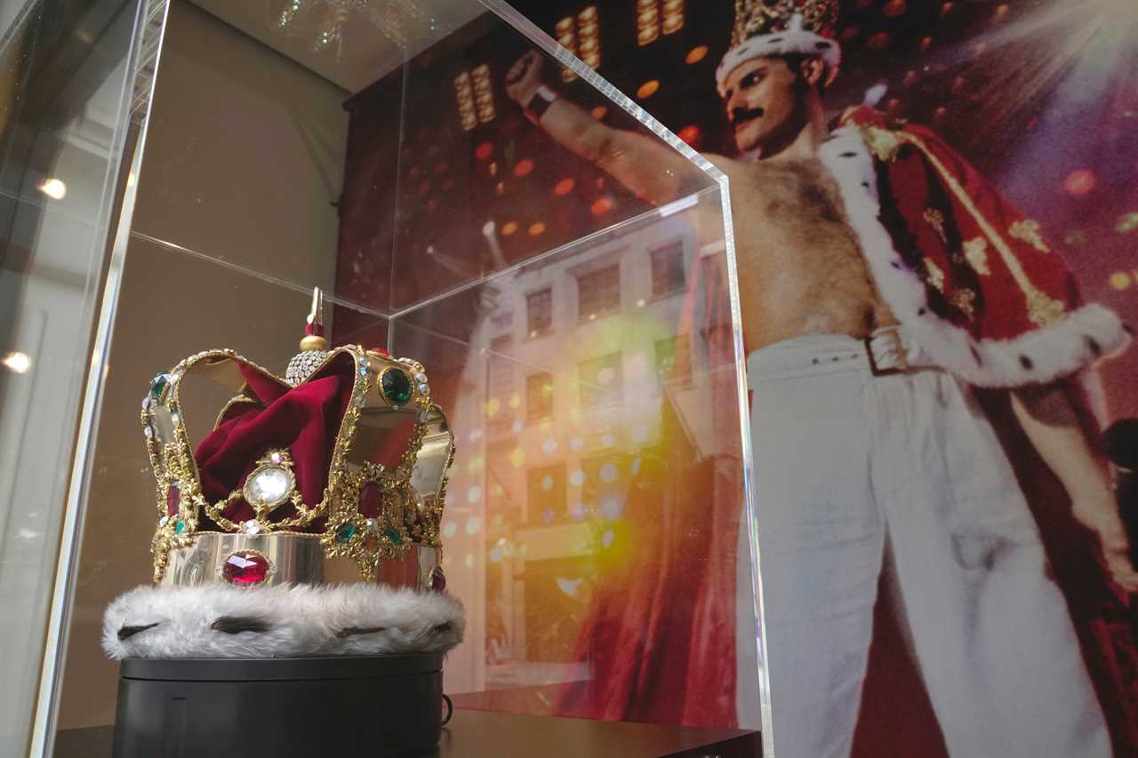 A red and gold bejeweled crown in a glass case in front of a large picture of Freddie Mercury.