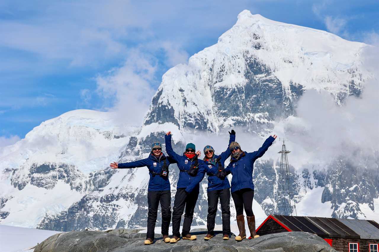 four women, Clare Ballantyne, Mairi Hilton, Natalie Corbett, and Lucy Bruzzone, pose with arms outstretched in front of a snowy mountain in Port Lockroy, Antarctica