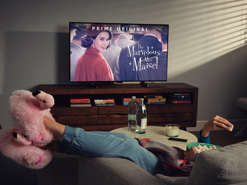 A person sits on a couch while eating popcorn and watching "The Marvelous Mrs. Maisel" on a TV.