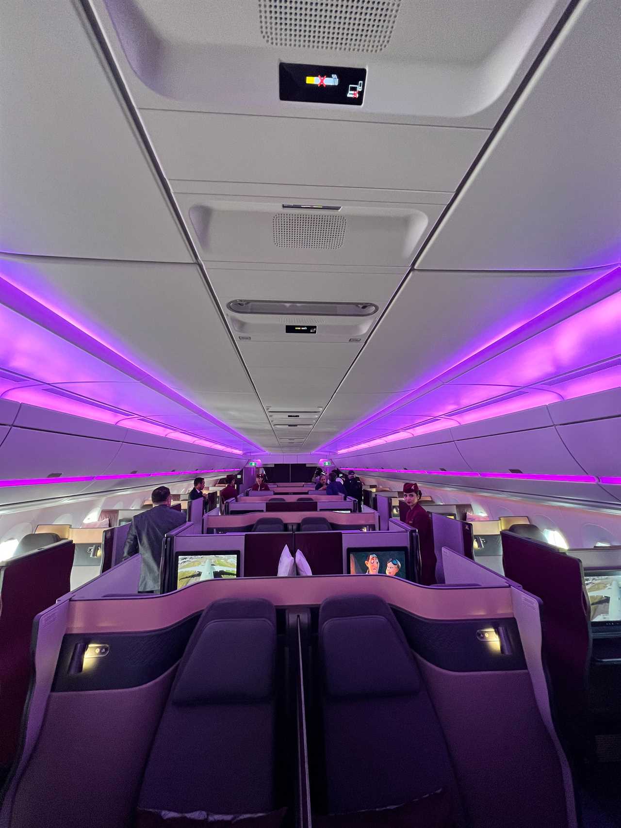 The QSuite business class cabin with purple accent lighting above the stowage bins, flight attendants in the Qatar Airways uniform and journalists walk down the aisles