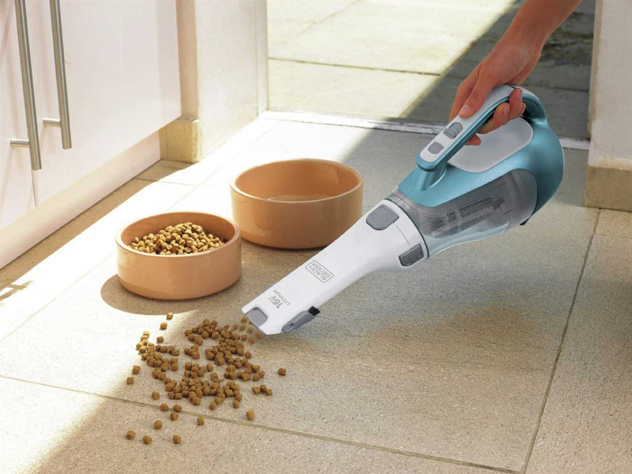 A person uses a BLACK+DECKER Dustbuster to clean up pet food around a bowl.