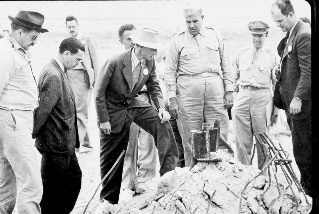 nine men in suits or beige army uniforms stand around a pile of wreckage black and white photo