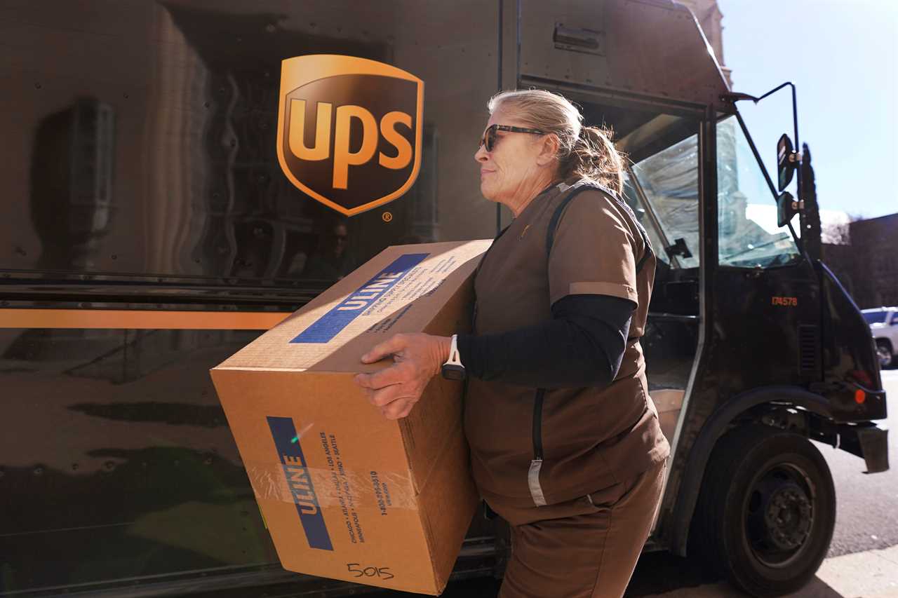 A blond woman in a brown UPS uniform carries a box in front of a brown UPS truck