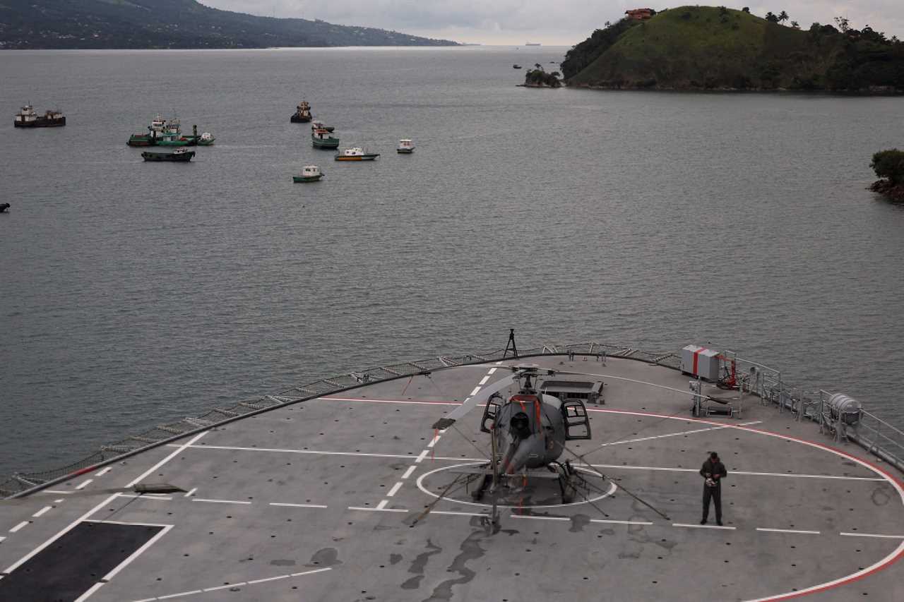 A man stands in the front of a Brazilian military ship, with a helicopter beside him, under a grey sky