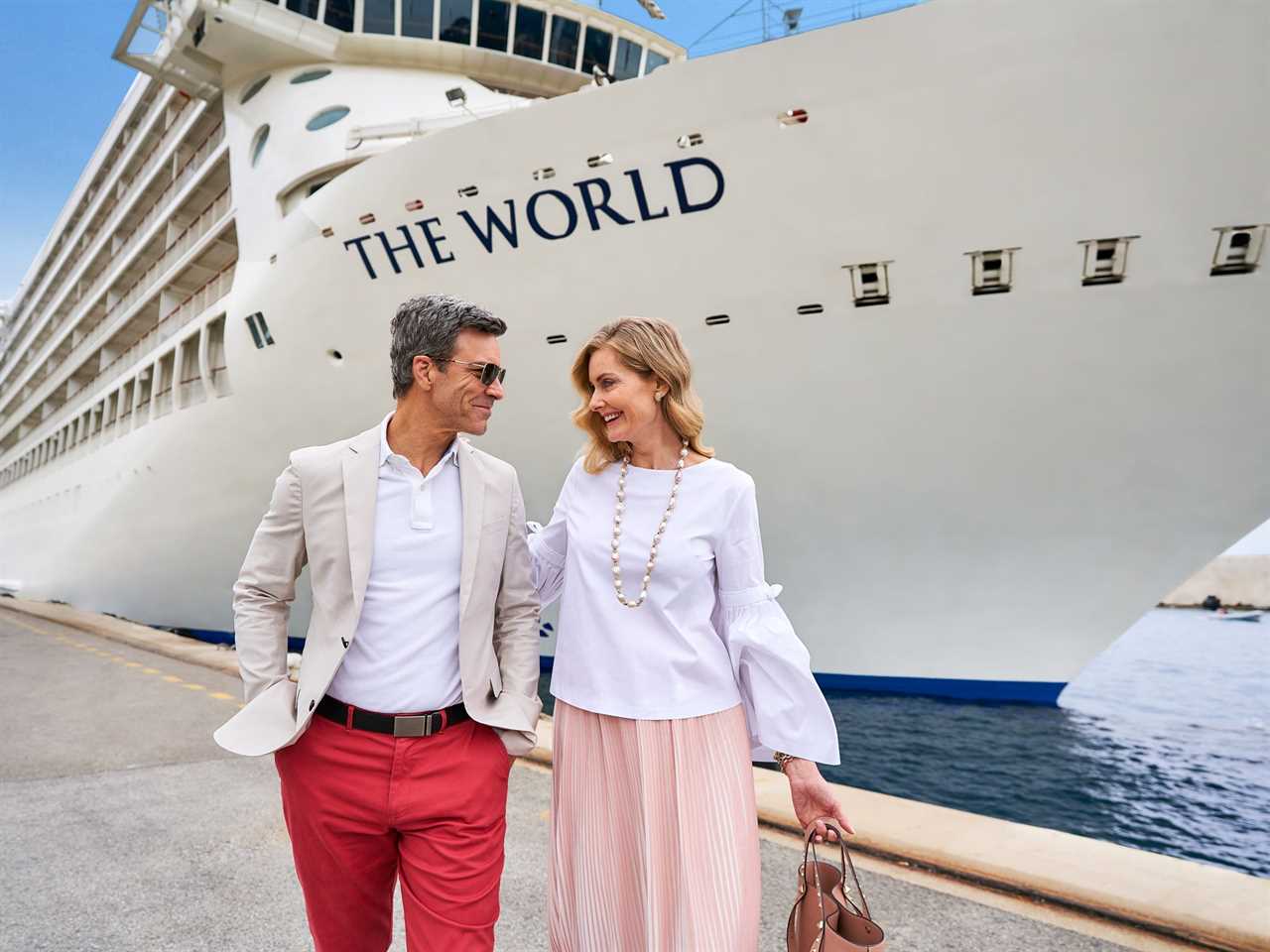 A couple smiling at each other while walking along a dock past the bow of The World.